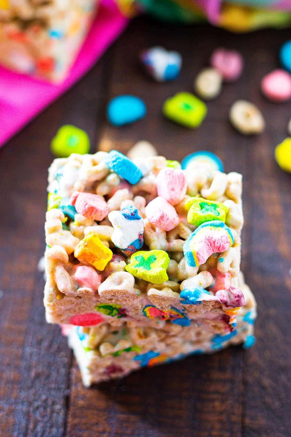 Lucky Charms Marshmallow Treats on a cutting board with Lucky Charms cereal scattered in the background.