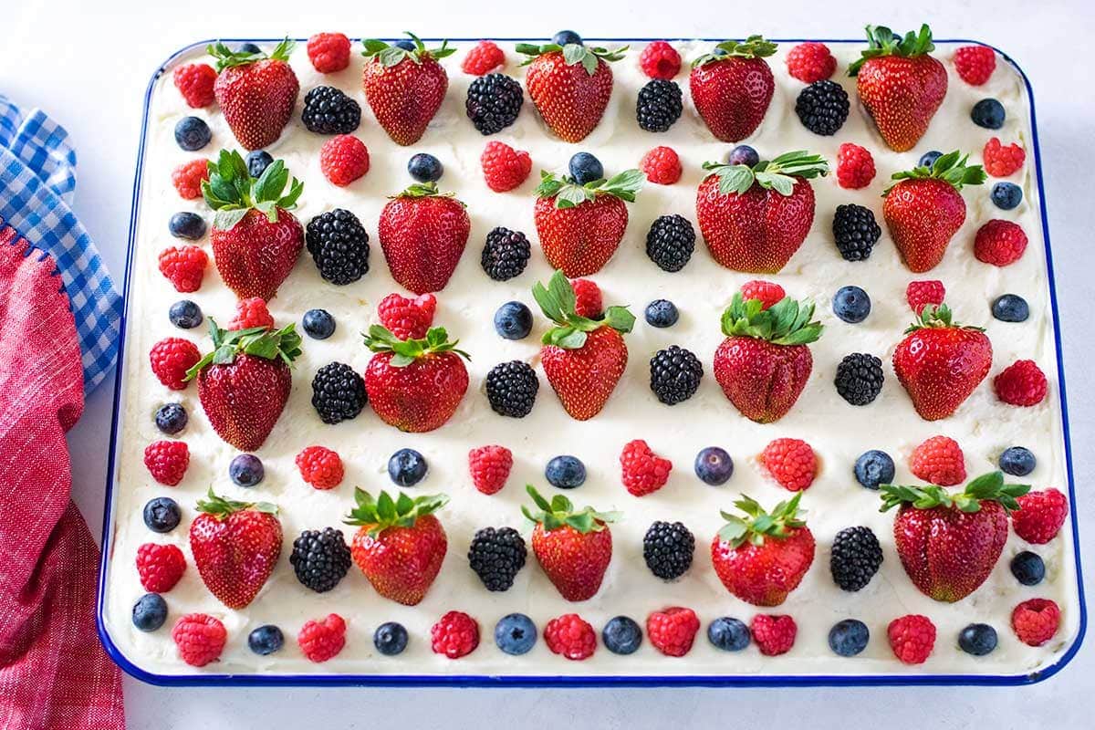 Chantilly sheet cake topped with fresh berries in a white enamel jelly roll pan with bright blue trim. Set on a white table with red napkin to the side.