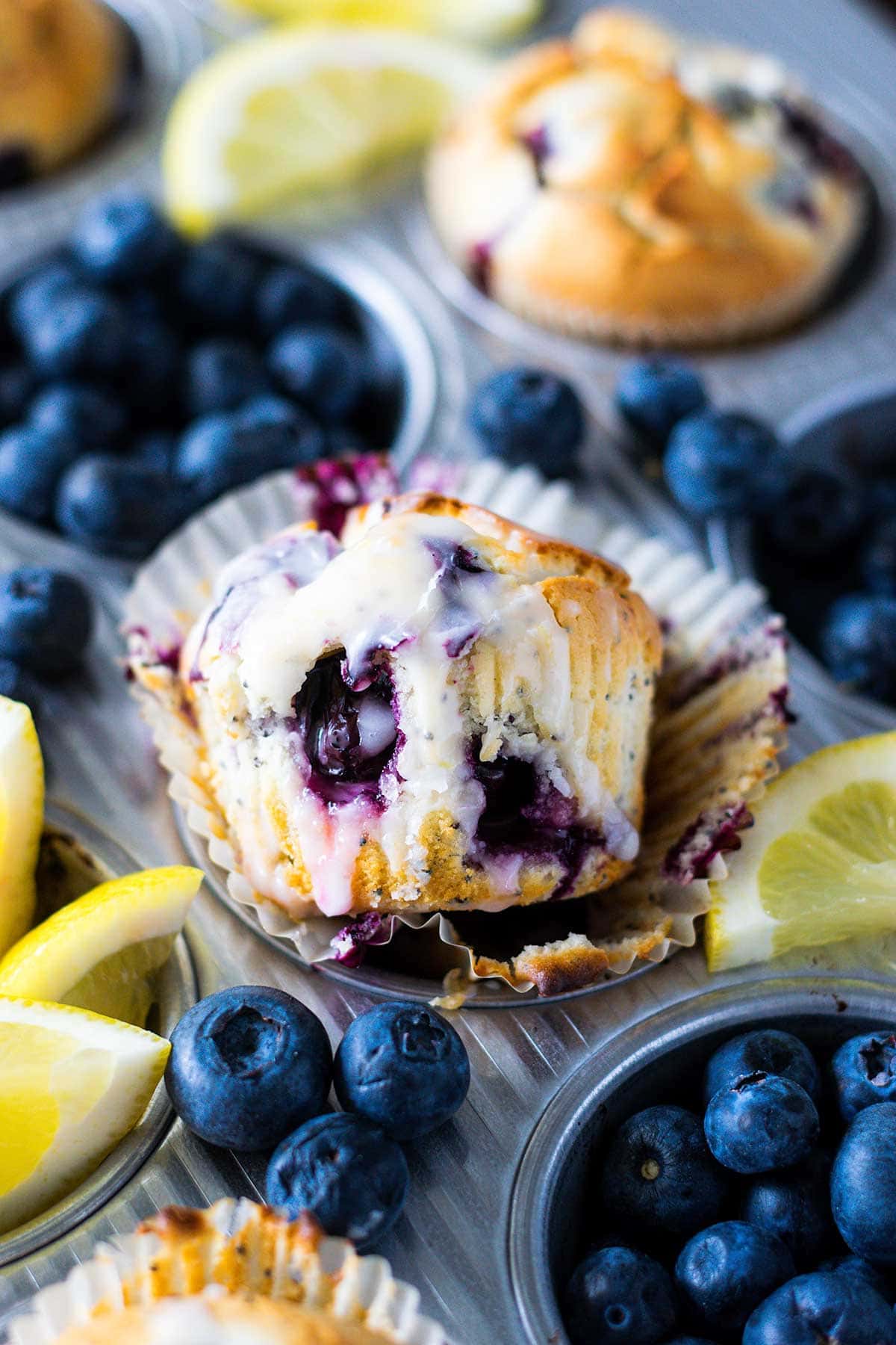 Blueberry lemon poppy seed muffin with lemon glaze drizzled on top in a muffin ray with scattered blueberries and lemon slices.