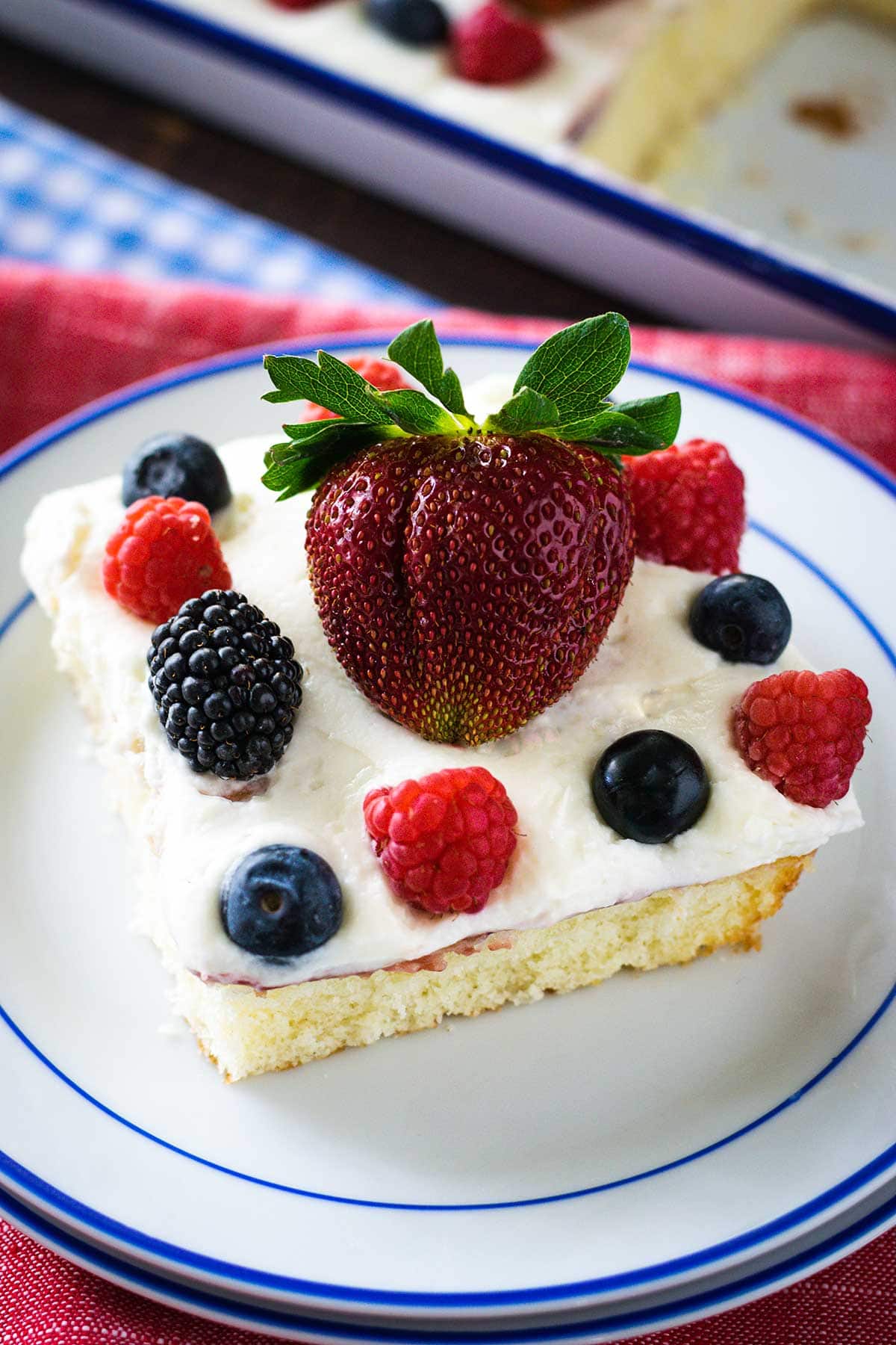 Up close image of a slice of chantilly cake topped with fresh berries on a white plate with blue trim. 