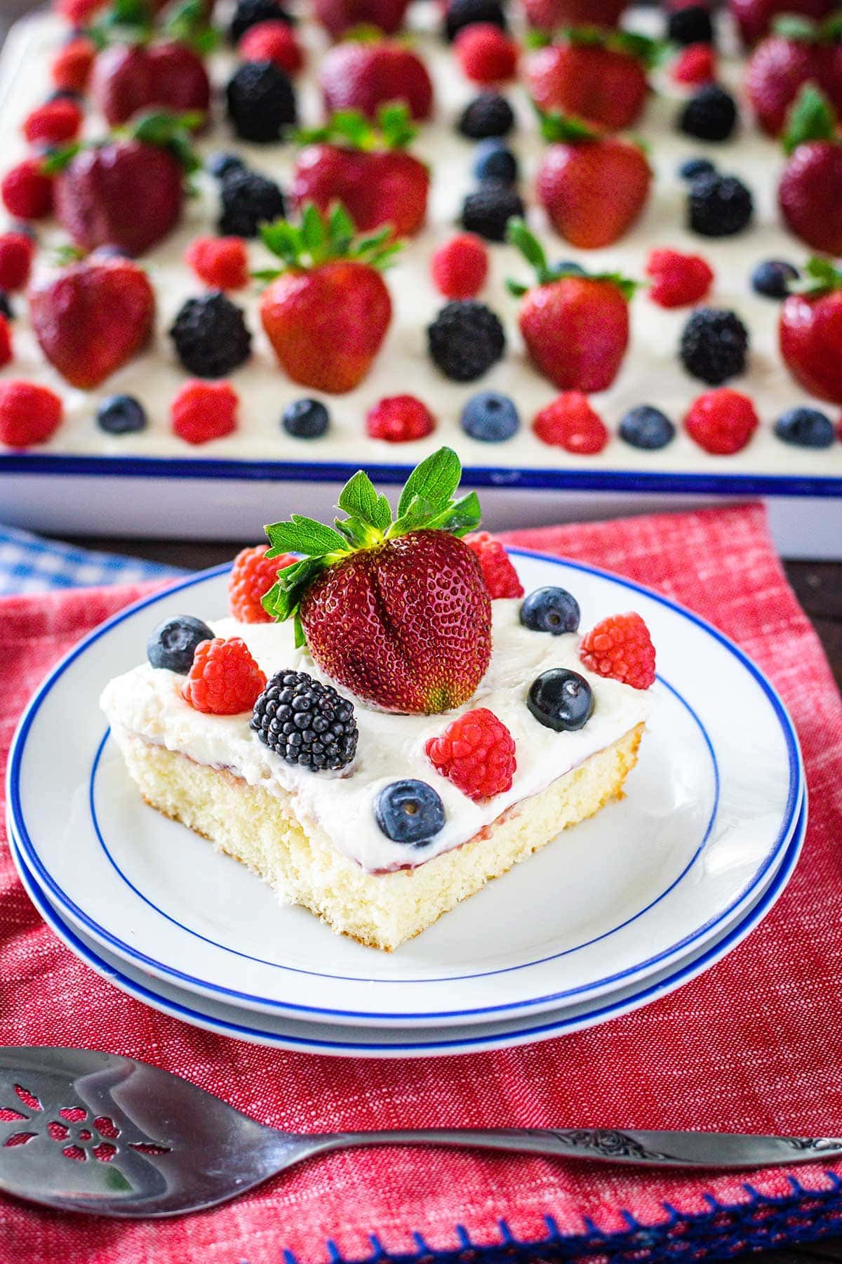 Slice of chantilly cake topped with fresh berries on a white plate with blue trim. Set on a red napkin with sheet cake in the background. 