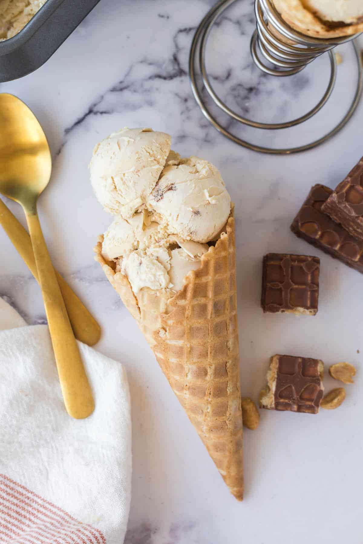 A waffle cone filled with nutty buddy ice cream on a marble table with nutty buddies to one side and two gold spoons on the other side.