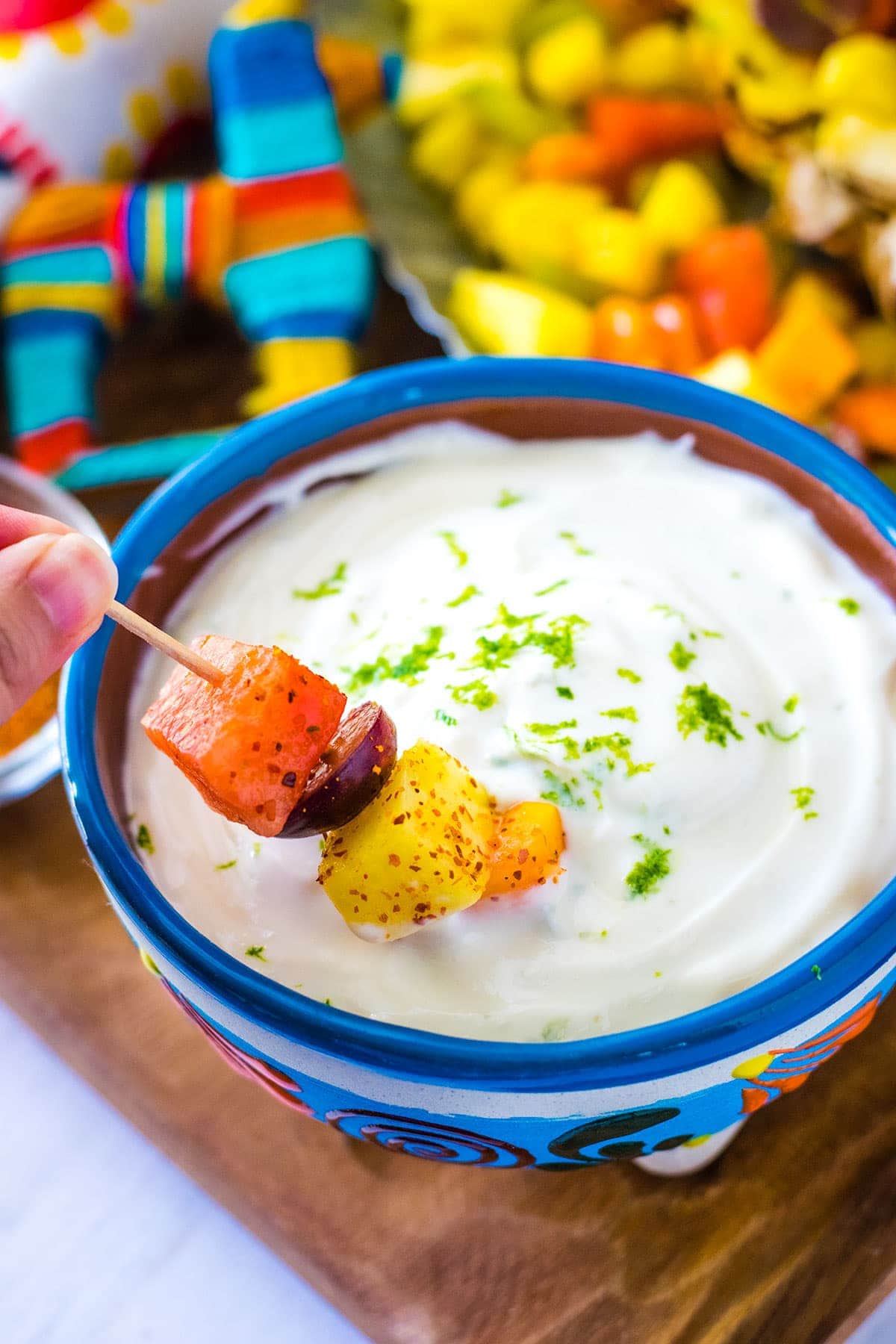 A fruit kabob being dipped in sweet lime yogurt dip. The dip is in a brightly colored bowl with a blue rim set on a wooden tray.
