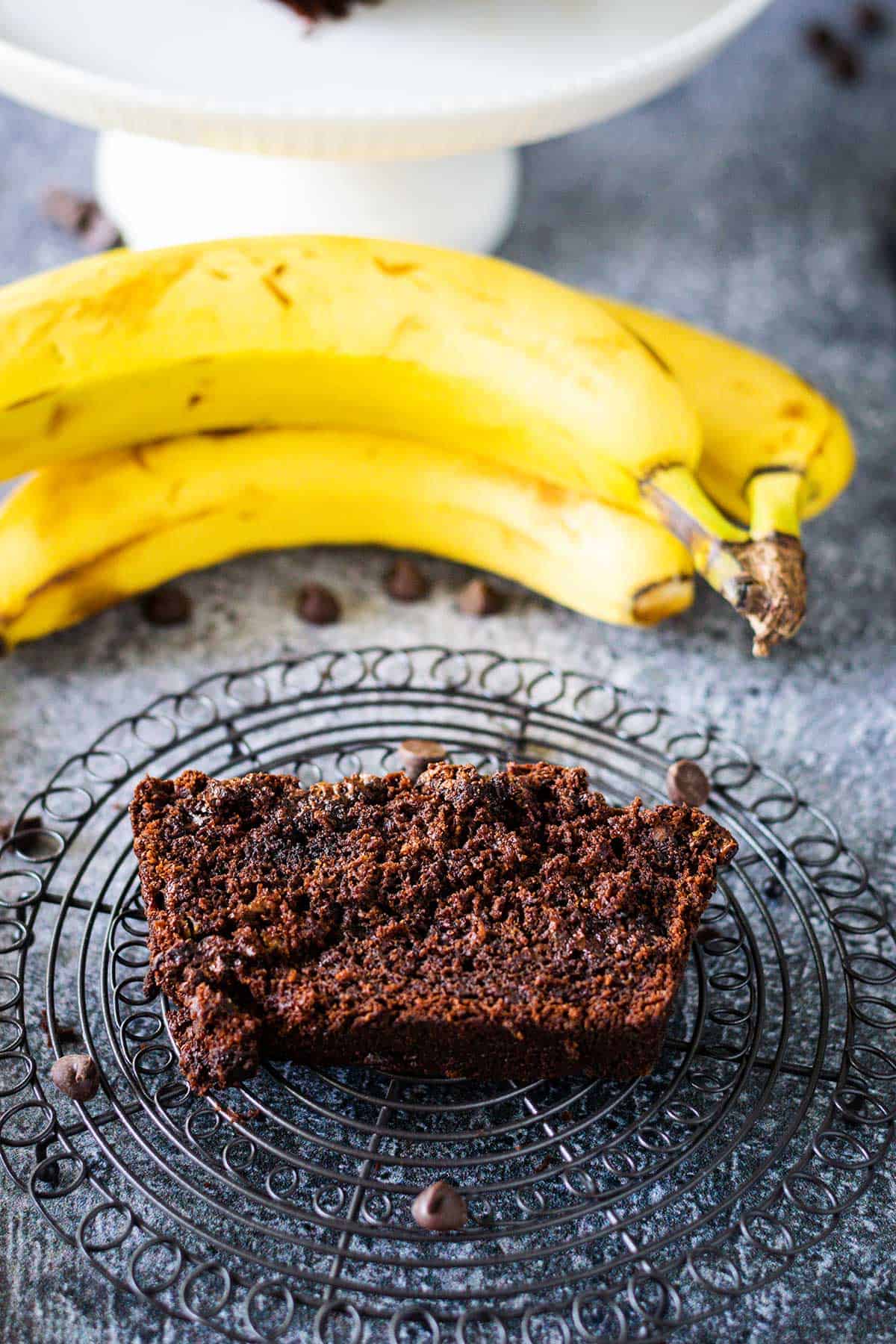 Slice of chocolate banana bread on a decorative round cooling wire with chocolate chips scattered and a banana in the background.