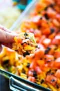 A Dish o mexican layer dip in the background with a person holding a chip filled with the dip.