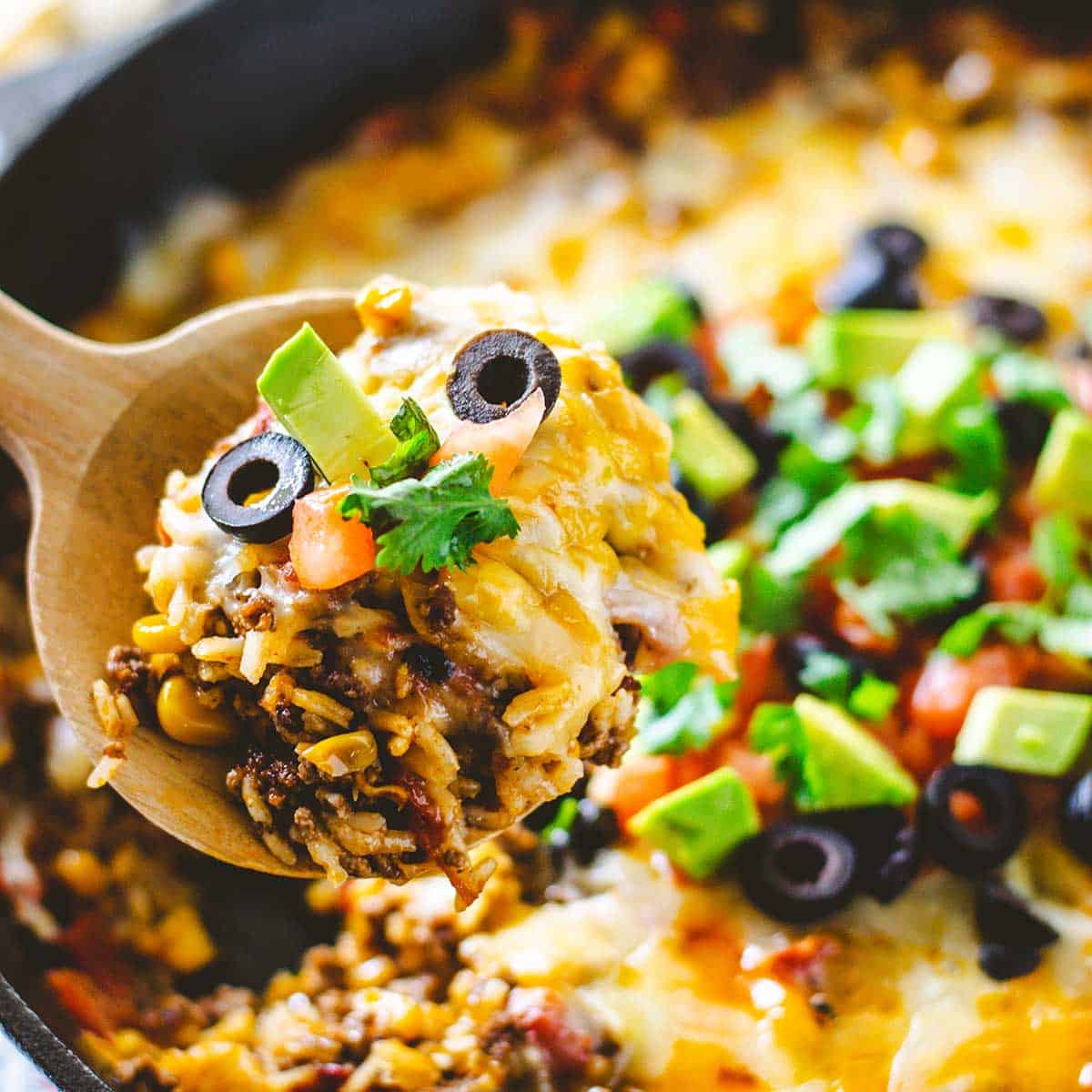 Upclose image of a scoop of ground beef and rice with cheese, avocado, black olives and tomatoes on top.