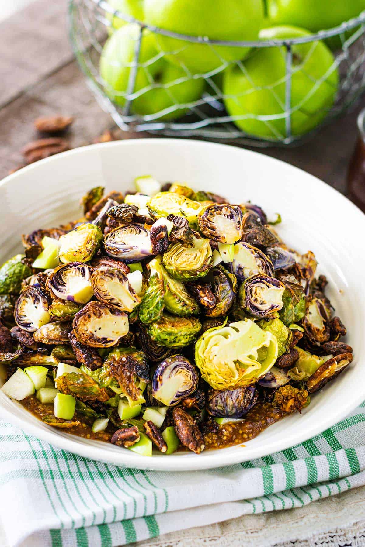 Plated apple butter dijon brussels sprouts served in a white bowl on a wooden table with a basket of green apples in the background.