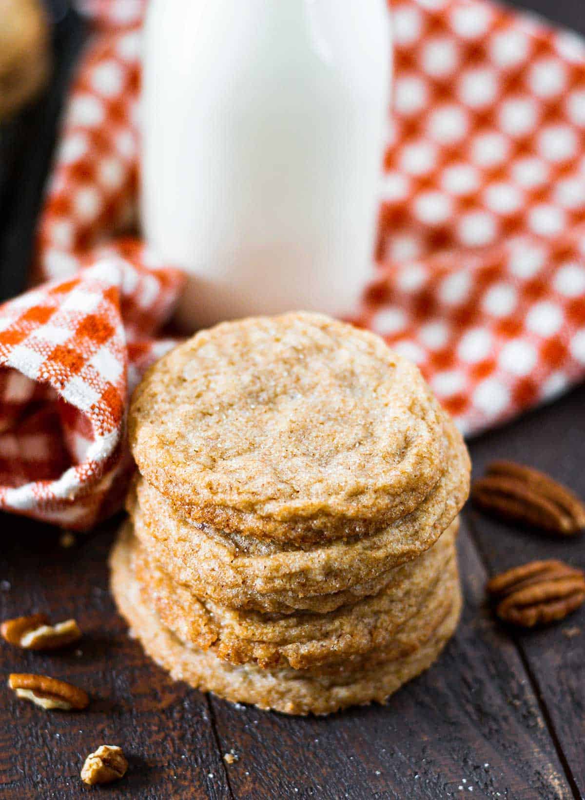 Stacked soft chewy pecan butter cookies on a wooden tray with scatter pecans and sugar and milk in background.