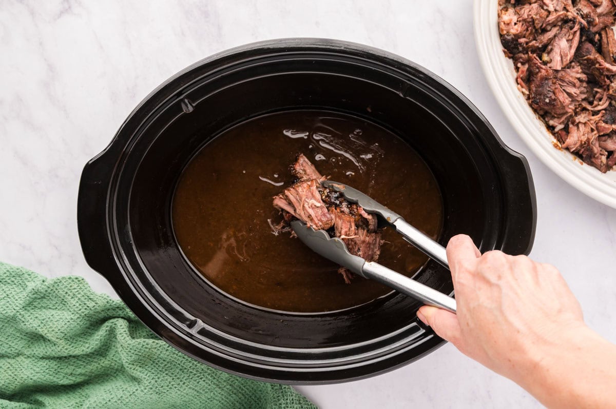 Using tongs to add the 3 packet pot roast back into gravy in the slow cooker.
