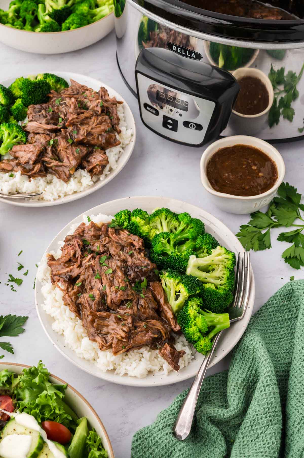 Two white plates with servings of 3 packet pot roast and gravy over white rice with broccoli served on the side with a gravy boat and the slow cooker in the background.