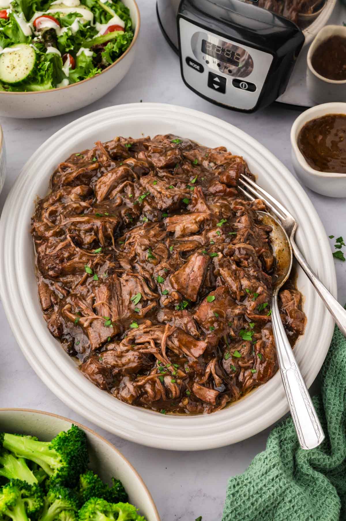 A white oval platter with shredded 3 packet pot roast and the slow cooker in the background.