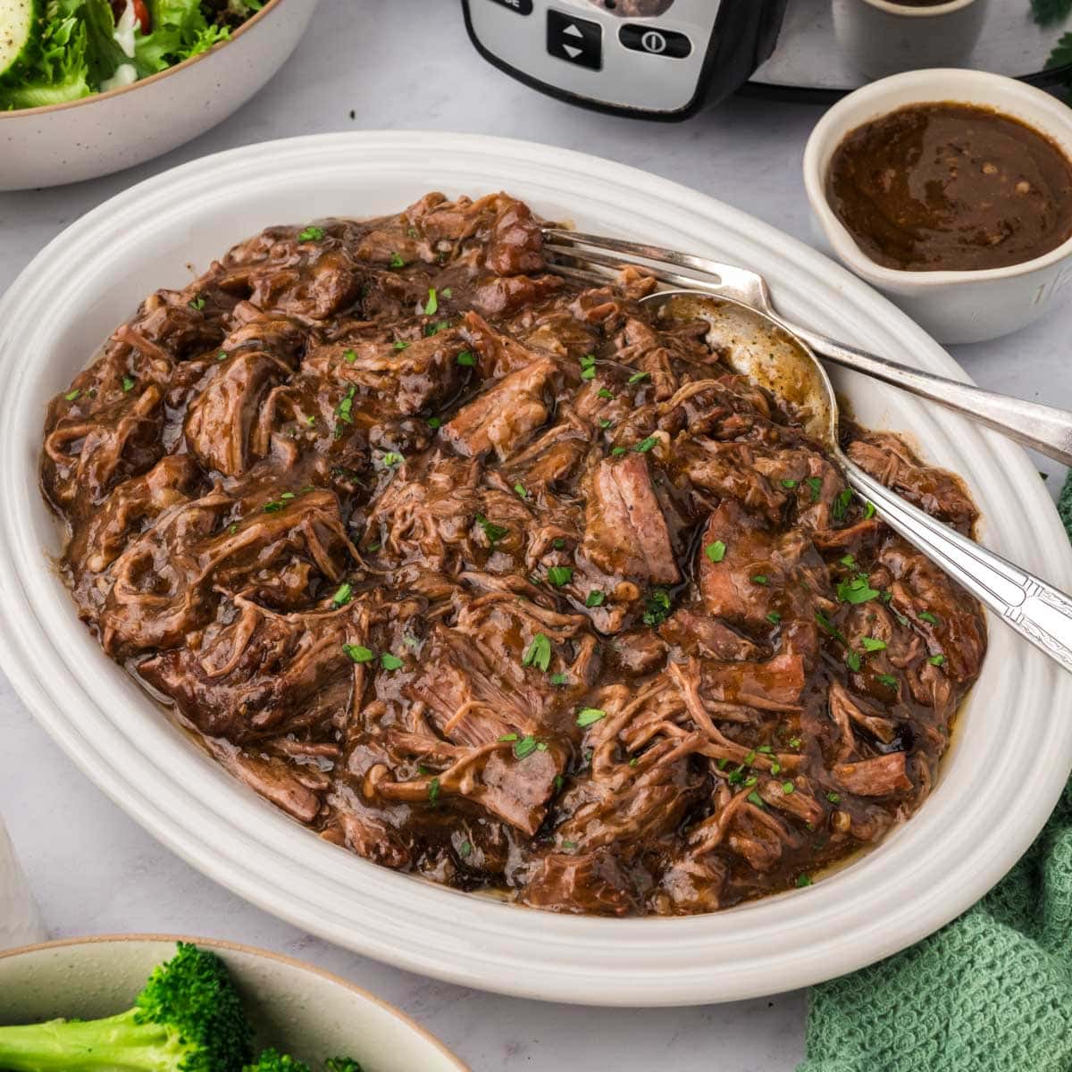A white oval serving platter of 3 packet pot roast and gravy with a gravy boat and the slow cooker in the background.