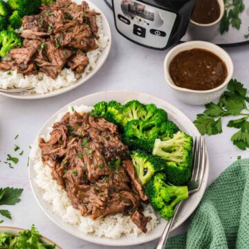 Two white plates with servings of 3 packet pot roast and gravy over white rice with broccoli served on the side with a gravy boat and the slow cooker in the background.