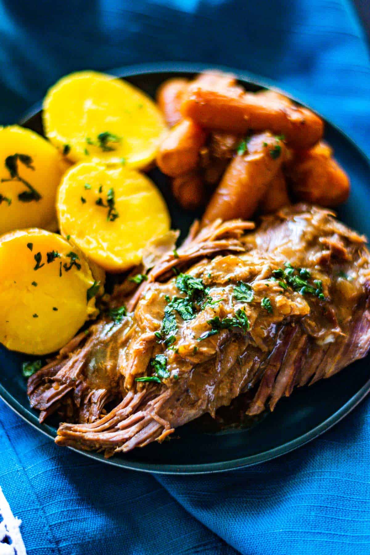 Slices pot roast with gravy on a blue plate with sliced potatoes and carrots.
