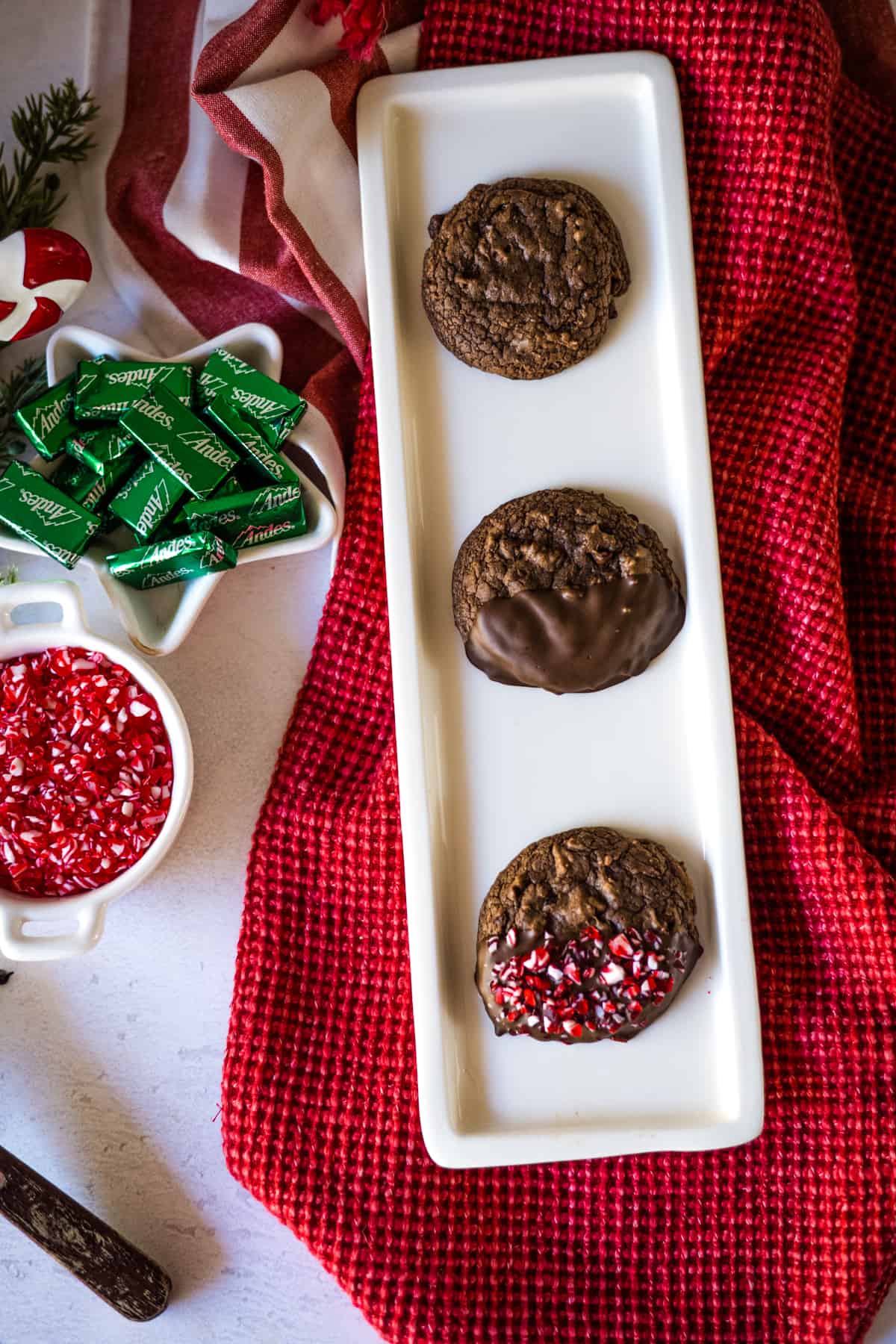 Peppermint Brownie Cookies on a narrow tray in stages of dipping the cookie, with sprinkles and Andes mints to the side on the table.
