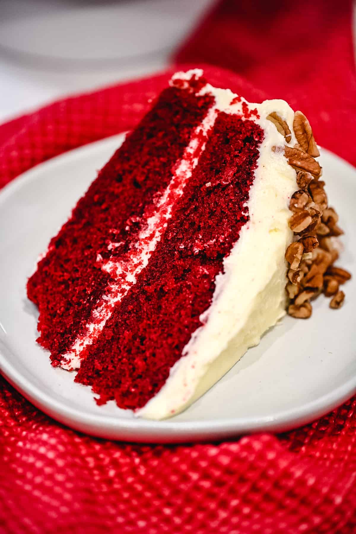 Slice of red velvet cake with cream cheese icing and pecans set on a red cloth.