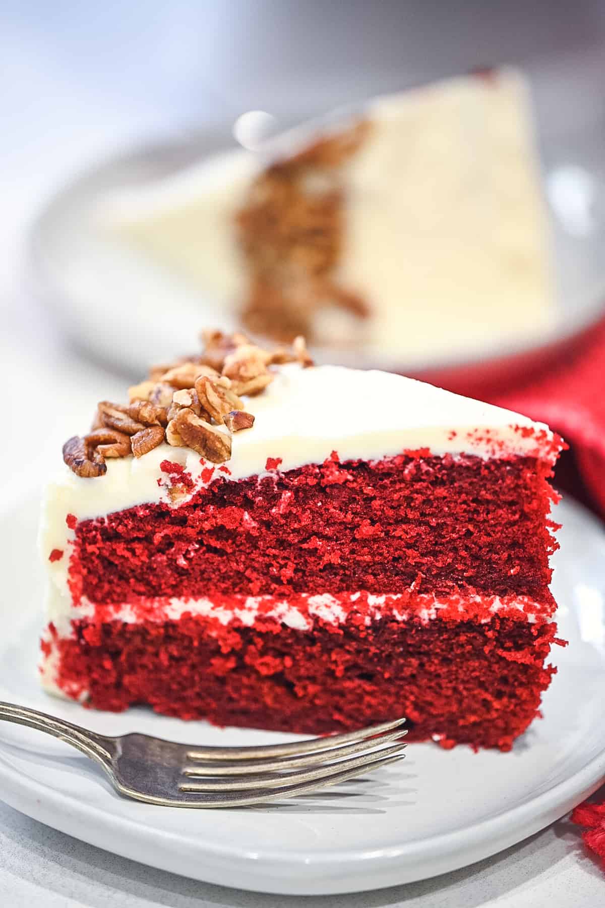 A upclose image of a slice of red velvet cake on a white plate with a fork in front of the cake.