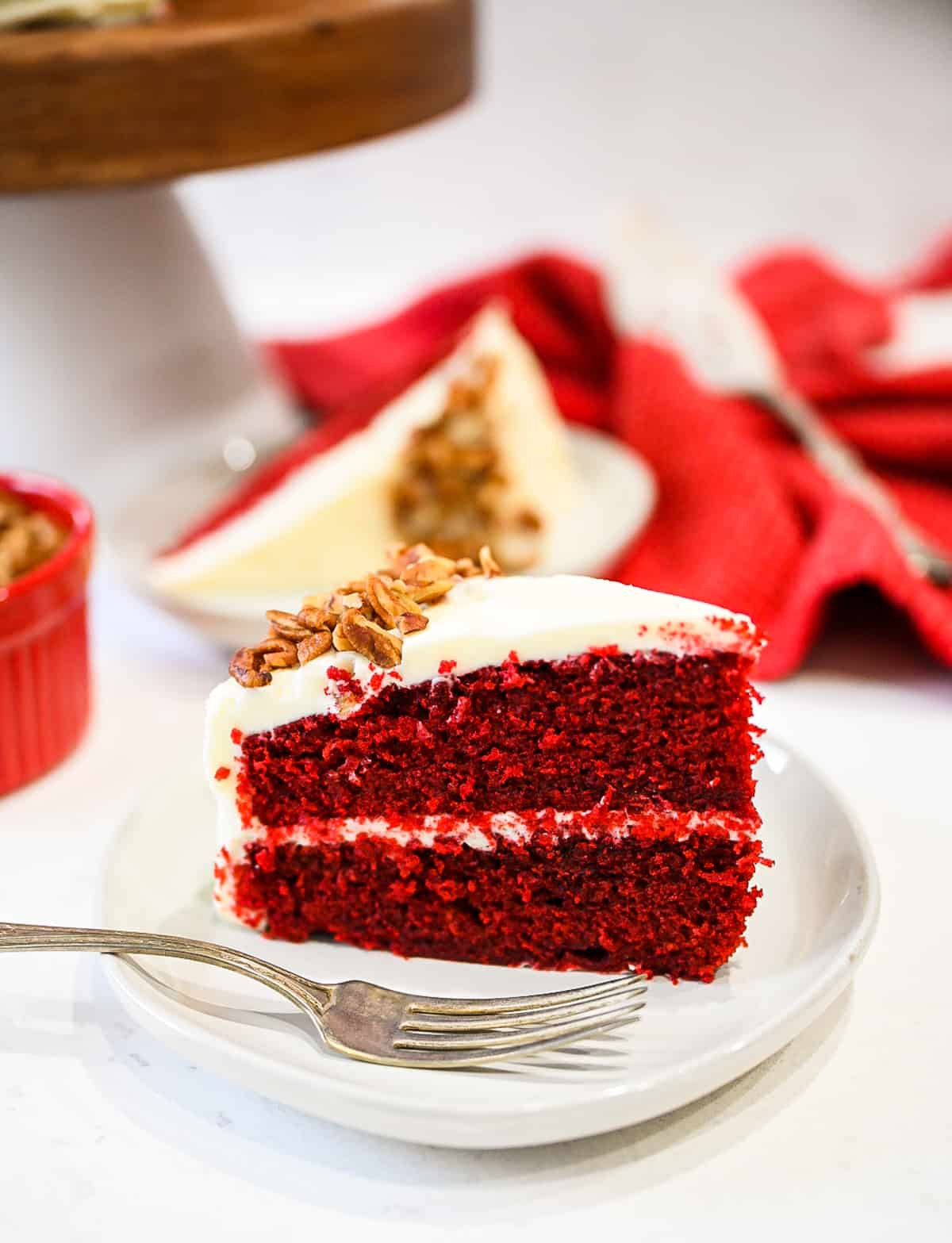 Up close image of classic red velvet cake with another slice in background set on a red cloth with cake server.