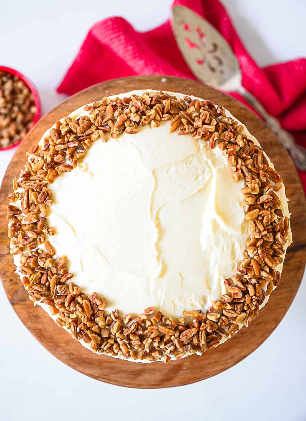 Overhead image of red velvet cake iced with cream cheese icing and garnished with chopped pecans on a wooden cake plate.