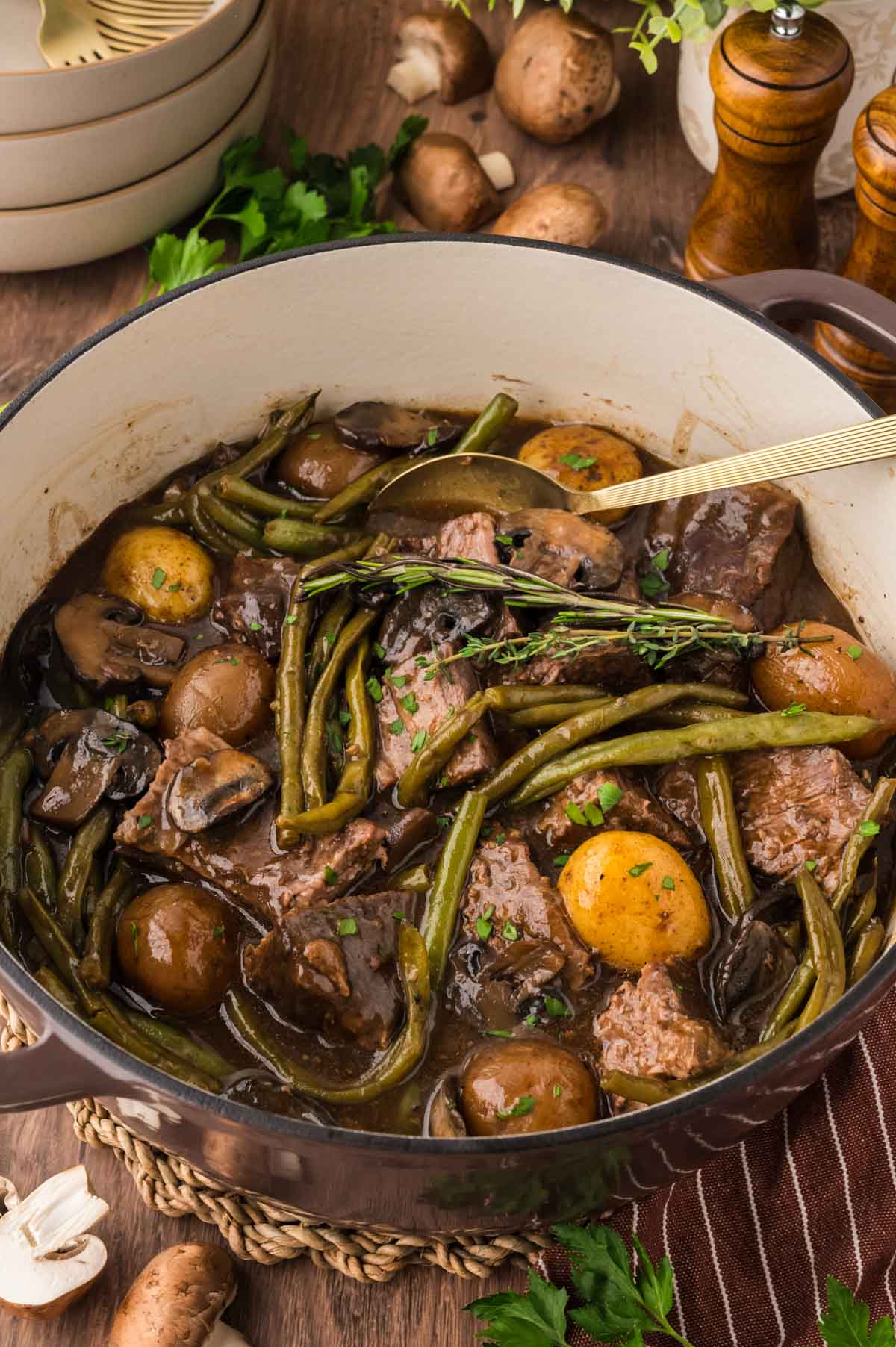 Pot roast, green beans, mushrooms, and potatoes in a brown dutch oven with a spoon serving spoon.