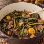 Pot roast, green beans, mushrooms, and potatoes in a brown dutch oven with a spoon serving spoon.