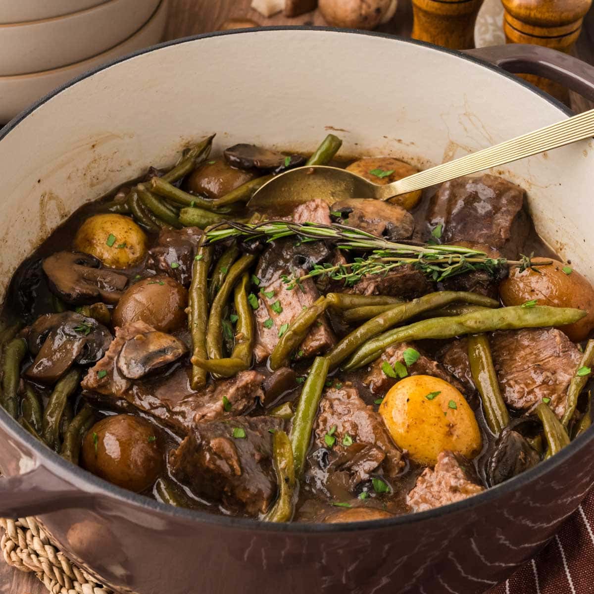 Pot roast, green beans, mushrooms, and potatoes in a brown dutch oven with a spoon serving spoon.