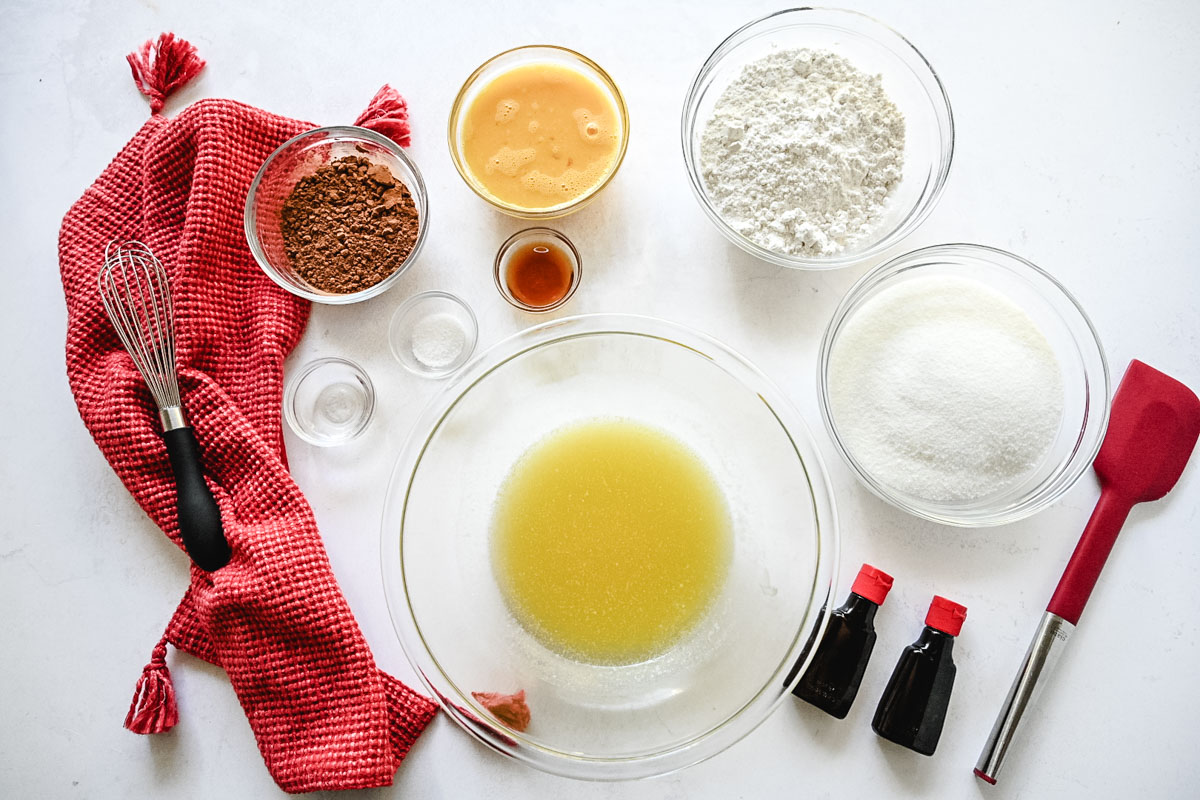 Image of red velvet brownie ingredients on a white tray with a red cloth napkin, a whisk, and a red spatula.