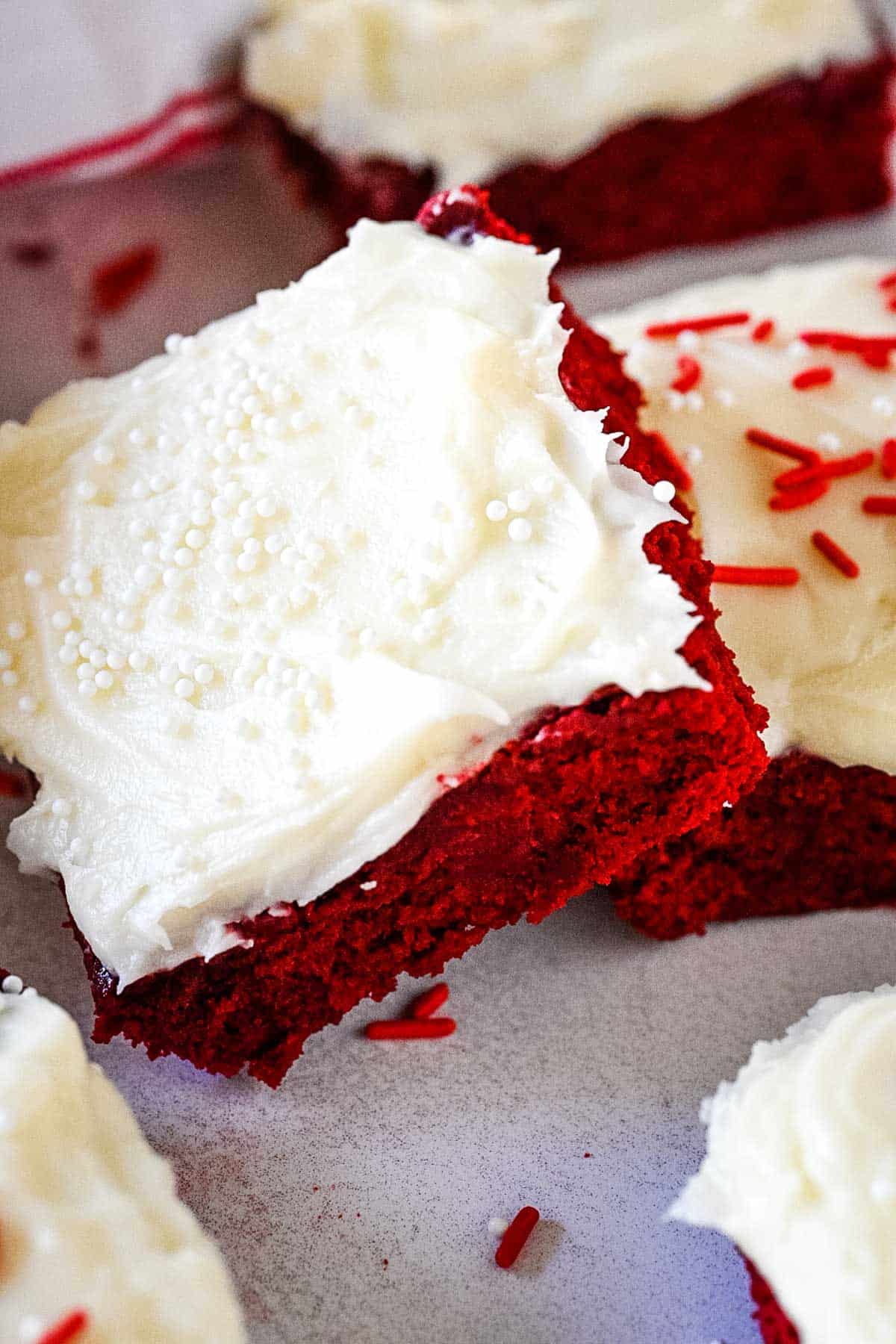 Red velvet brownie frosted with cream cheese icing and white sprinkles stacked at an angle on top of another brownie.