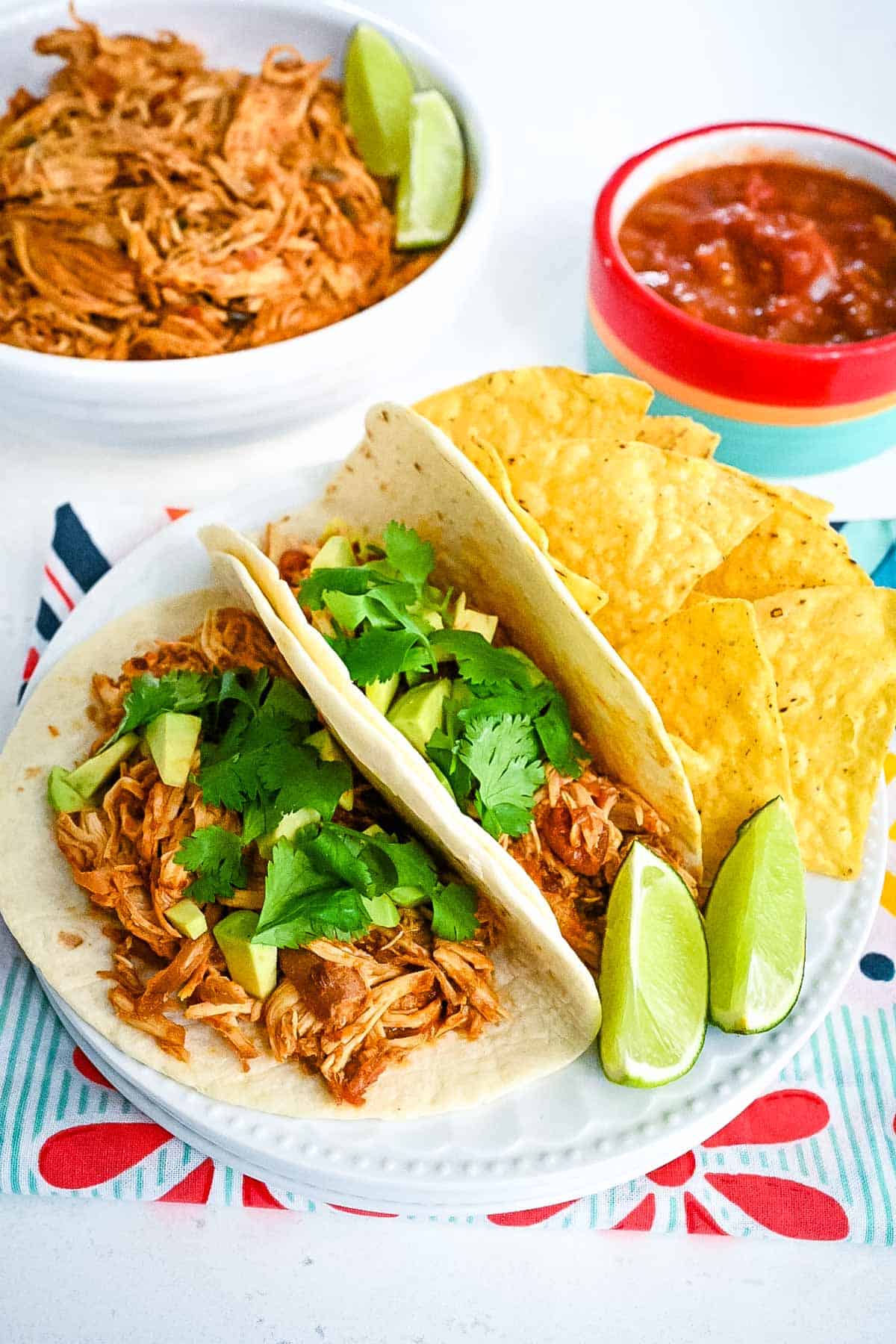 Soft tacos made with shredded salsa chicken on a white plate with tortilla chips and lime wedges.