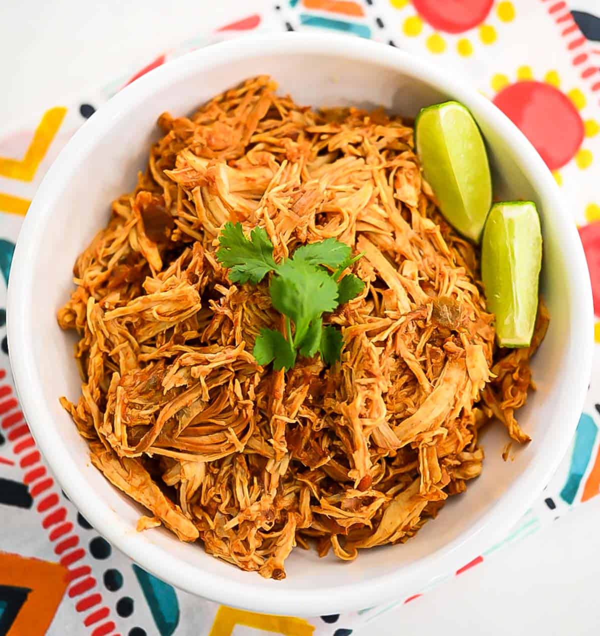 An overhead image of a white bowl filled with slow cooker salsa chicken that has been shredded and garnished with cilantro and lime wedges.