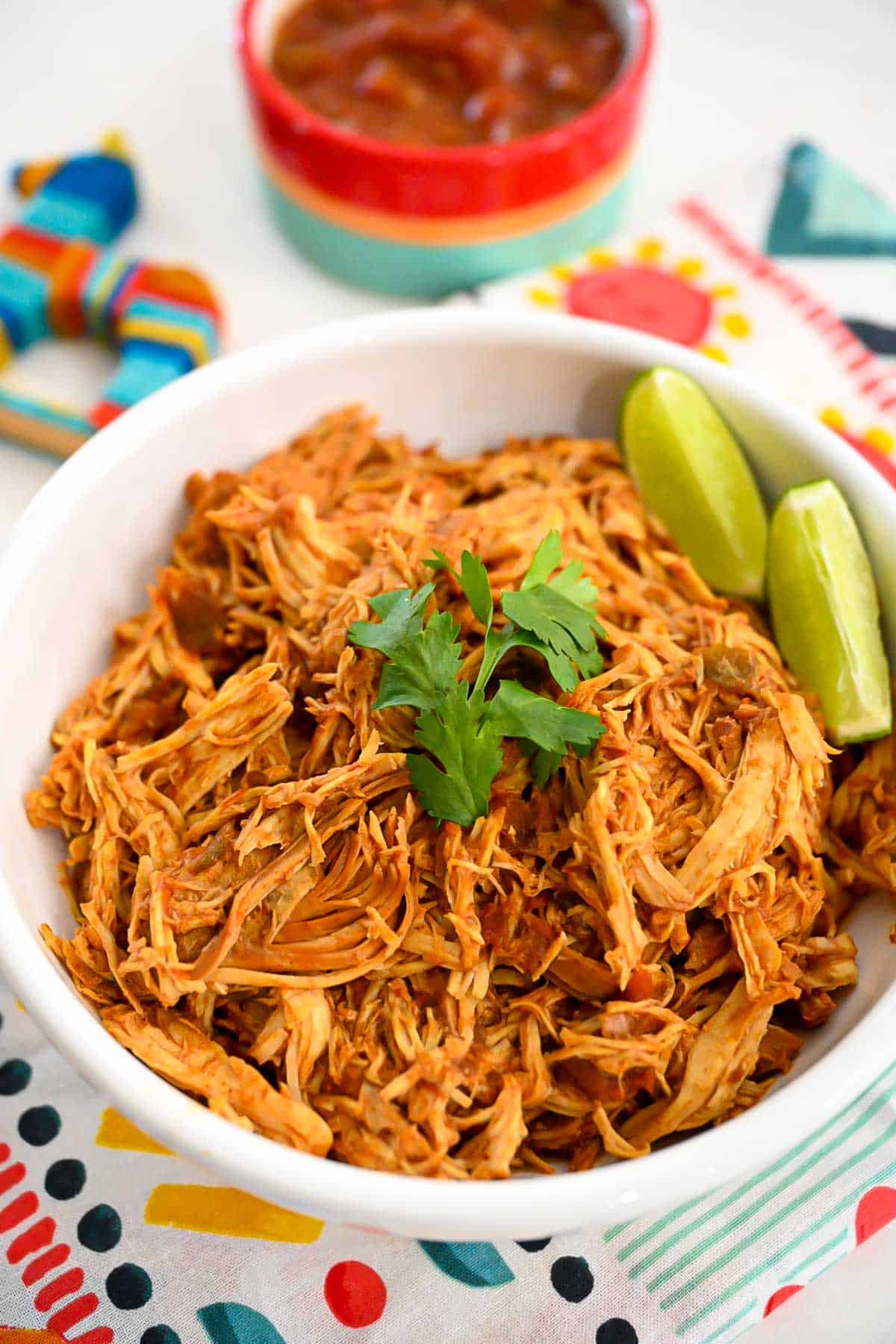Shredded slow cooker salsa chicken in a white on on a festive brightly colored table setting with a bowl of salsa in the background.