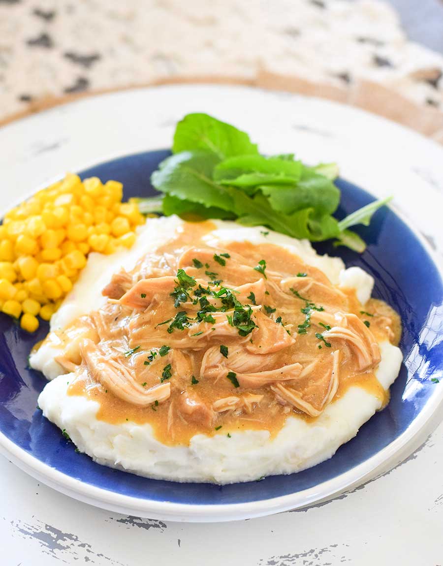 Plated slow cooker chicken and gravy over mashed potatoes. Served with corn and leafy greens.