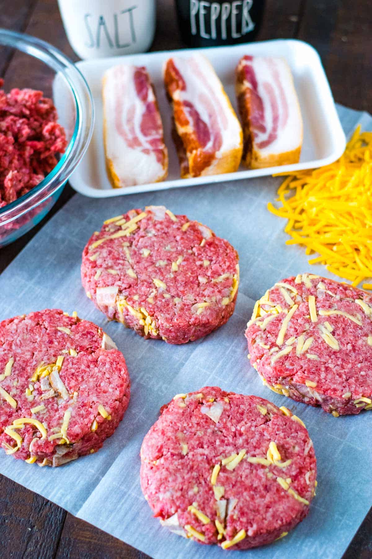 Stuffed bacon cheddar burger patties with all the ingredients needed to make in the background - ground round, bacon slices, cheddar cheese, salt, and pepper.