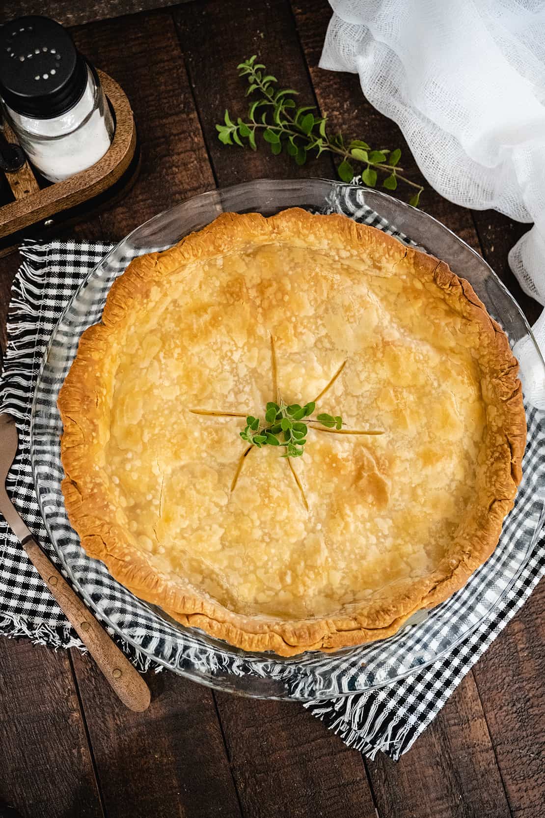 Overhead image of a whole chicken pot pie garnished with fresh thyme set on a wooden tablescape with a black and white checked placemat.