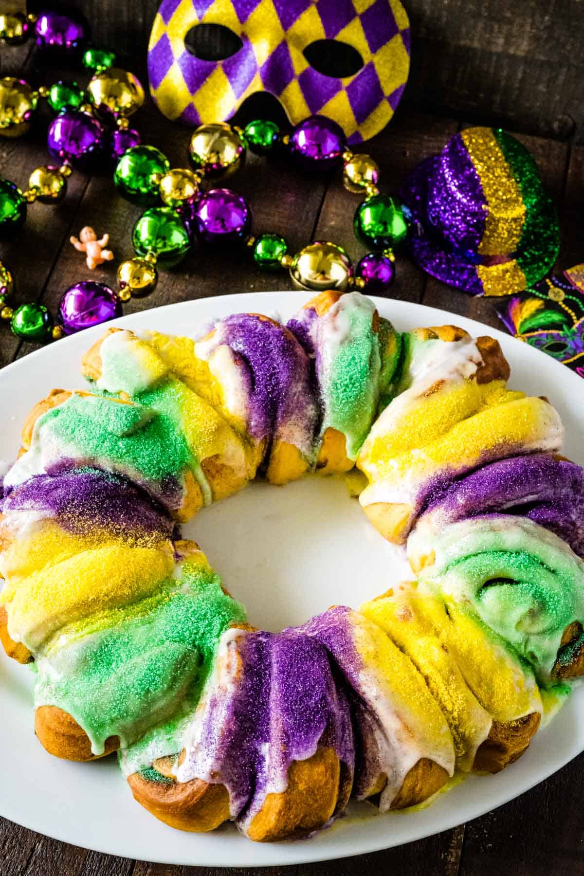 A king cake on a white oval platter set on a wooden table with mardi gras beads and a mask in the background for decoration.