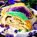 Slice of mardi gras king cake on a white serving plate with mardi gras beads scattered around the plate for decoration.