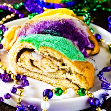 Slice of mardi gras king cake on a white serving plate with mardi gras beads scattered around the plate for decoration.