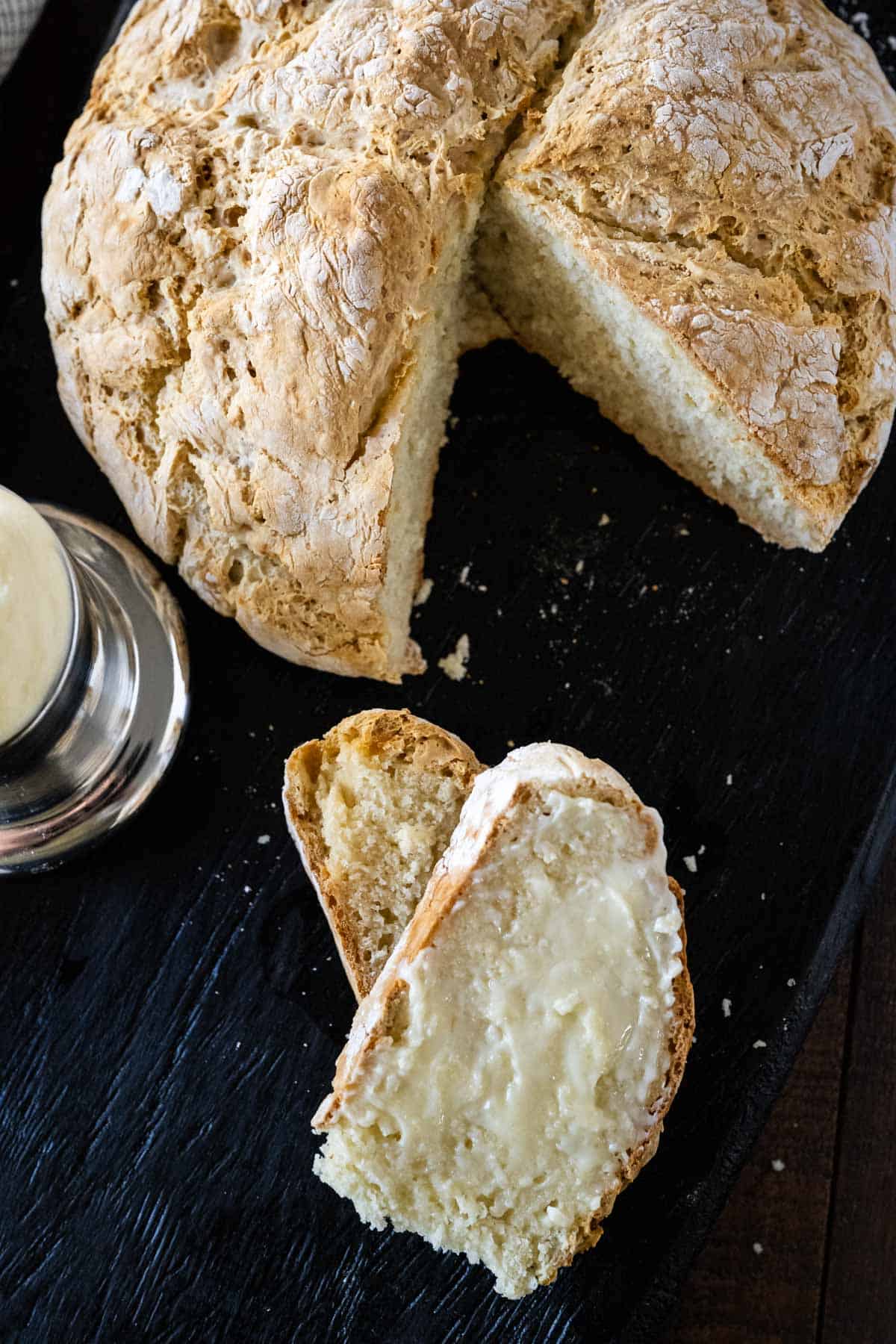 Soda bread sliced with butter slathered on top set on a black wooden tray with the loaf and butter crock.