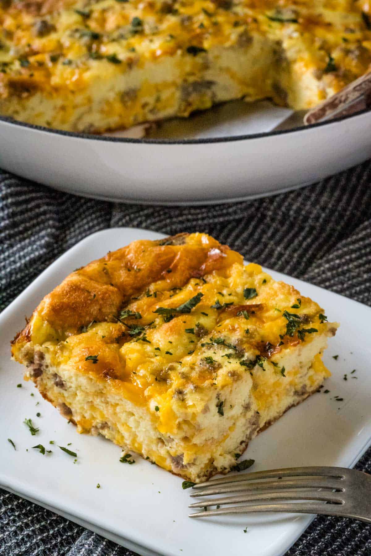 A slice of sausage, egg, and cheese casserole on a white plate with a fork in front of the slice.