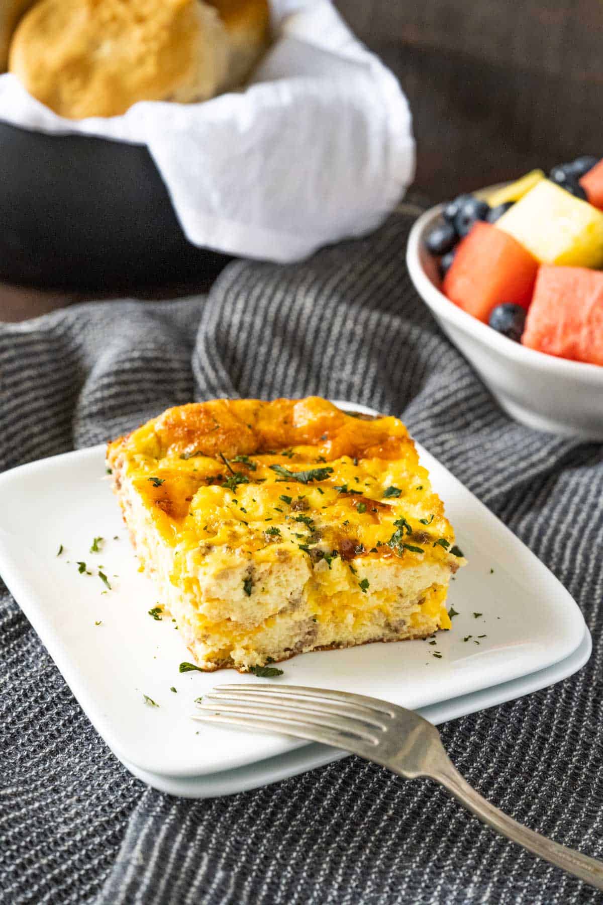 Fresh slice of breakfast casserole with a fruit bowl and biscuit basket in the background.