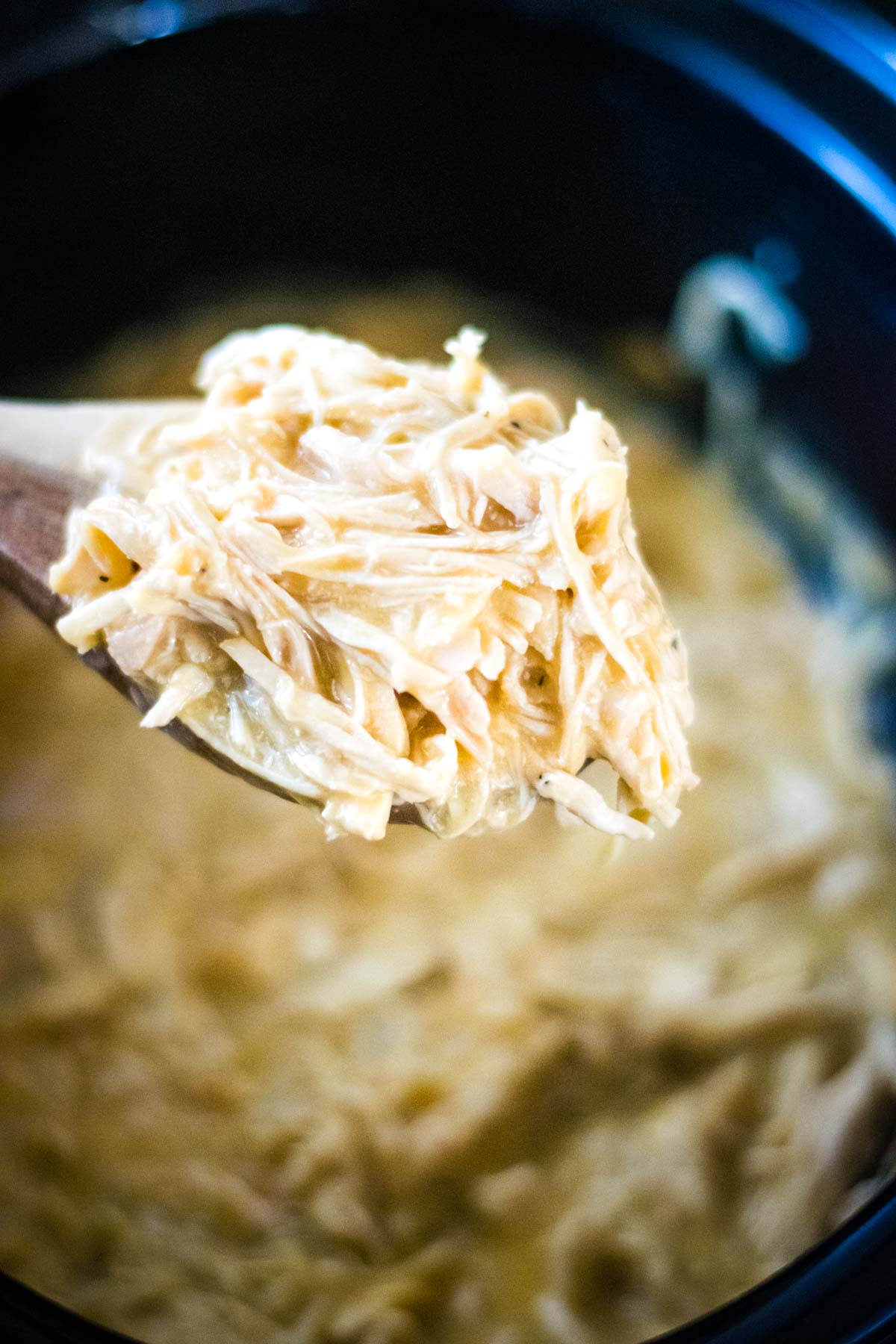 A spoon full of slow cooker chicken and gravy with the crock pot in the background.