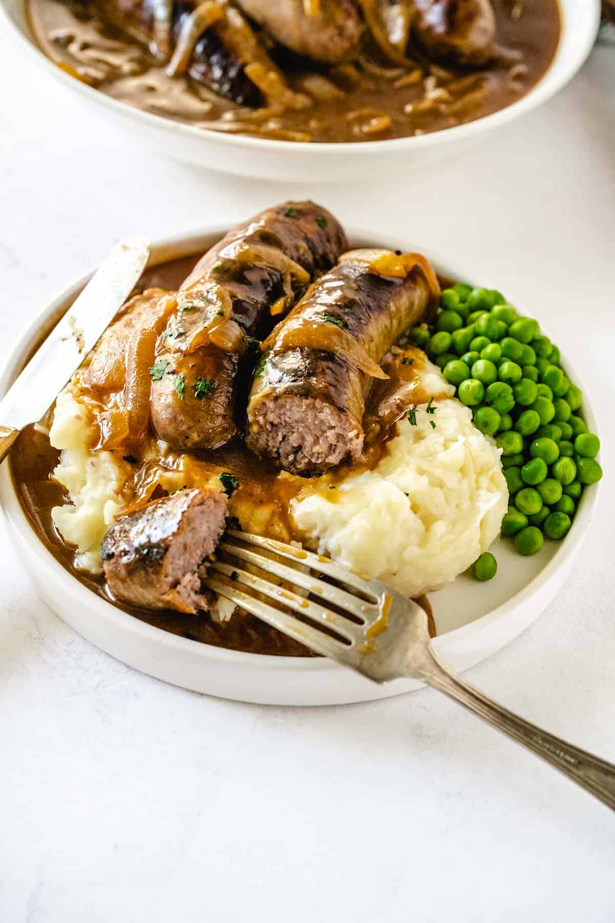 Bangers and mashed potatoes with gravy on a white plate with a fork and knife with one of the sausages is cut.
