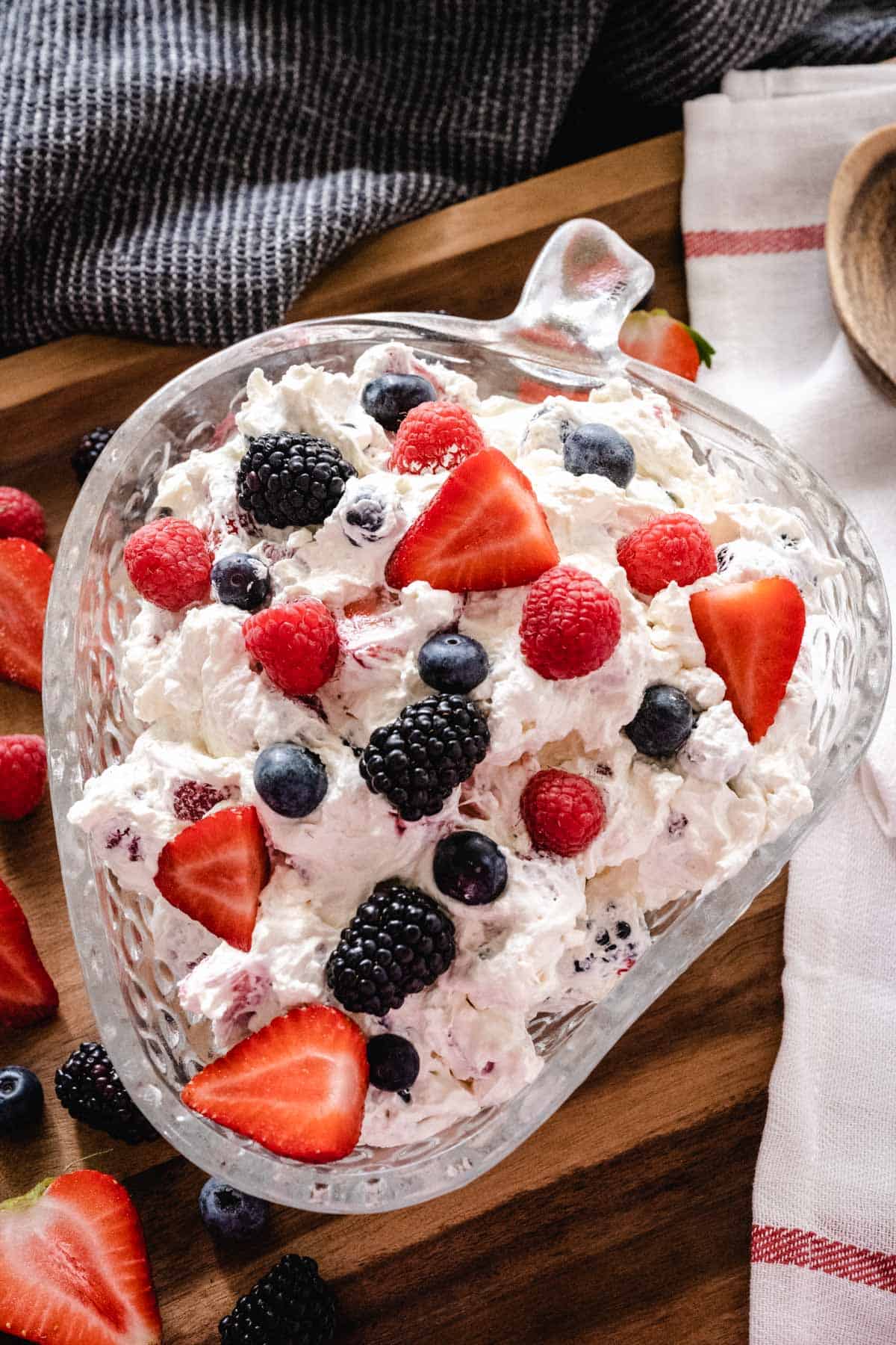 Berry cheesecake salad in a strawberry shaped bowl on a wooden tray with fresh cut berries scattered on tray.