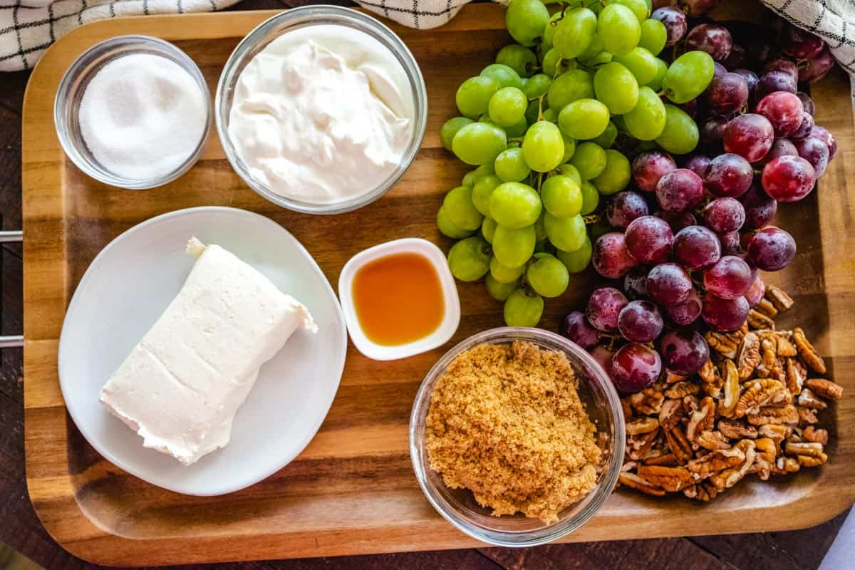 Image of ingredients needed to make grape salad recipe - grapes, cream cheese, sour cream, sugar, vanilla extract, brown sugar, and pecans.
