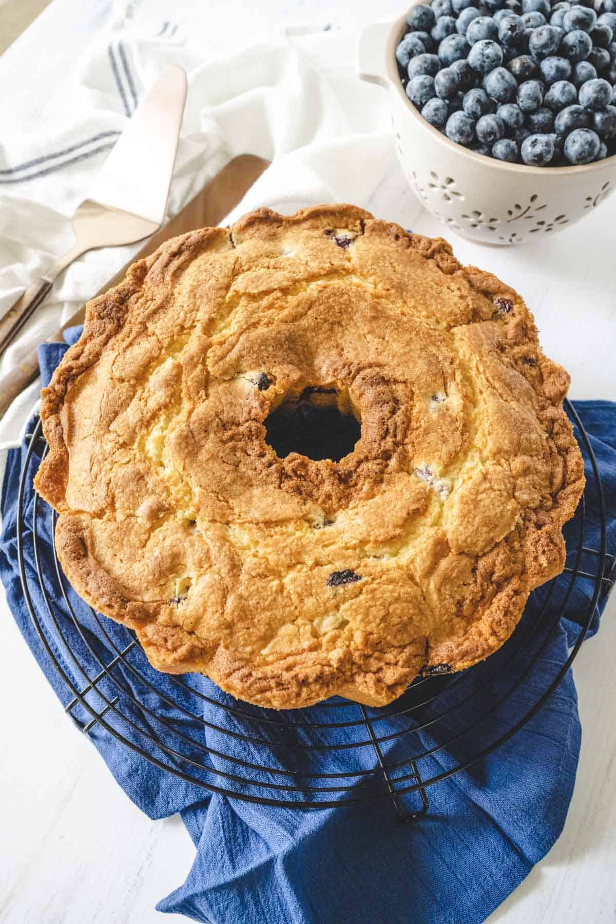 Overhead image of a whole blueberry pound cake on a blue table cloth with a bowl of blueberries in the backdrop.