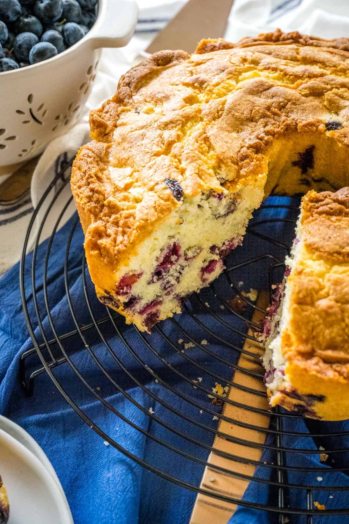 Blueberry Pound Cake with a slice removed on a wire cooling rack set on a blue cloth.