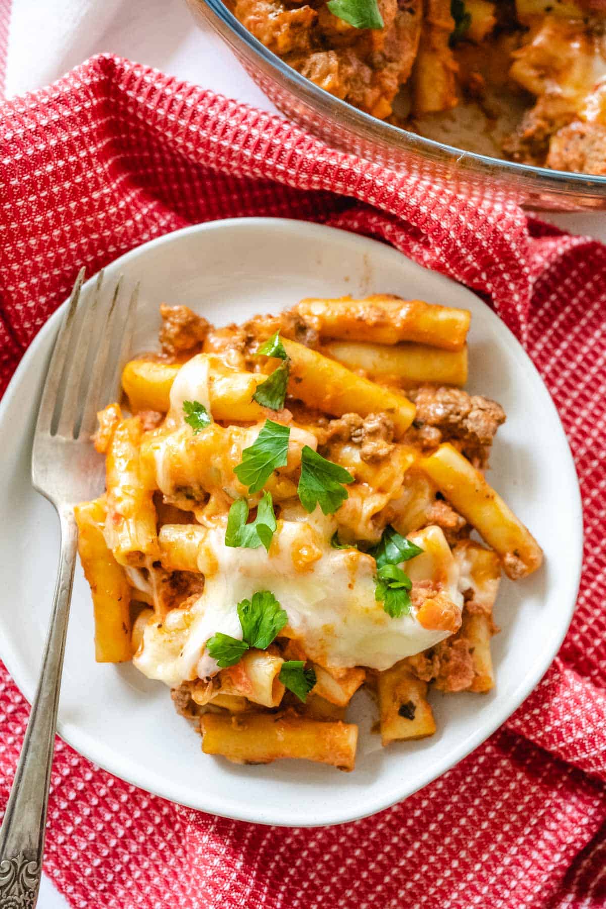 an overhead image of easy baked ziti placed on a white plate with a fork to the side with a red cloth underneath.