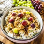 A big bowl of creamy grape salad on a wooden tray with grapes and pecans in the background.