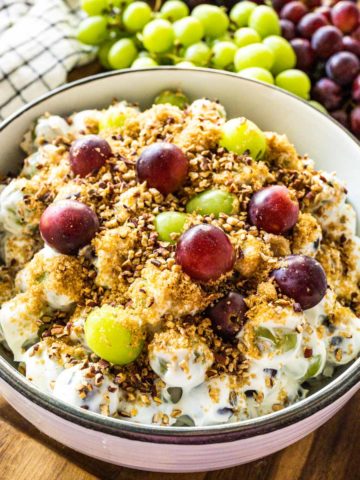 A big bowl of creamy grape salad on a wooden tray with grapes and pecans in the background.