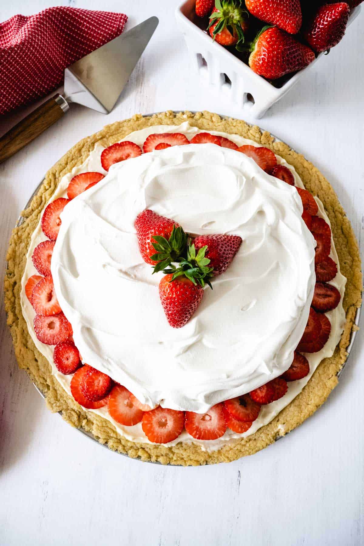 A strawberry pizza with a cookie dough crust, sliced strawberries and topped with whipped cream on a white wooden table with strawberries and a cake slicer in the background.